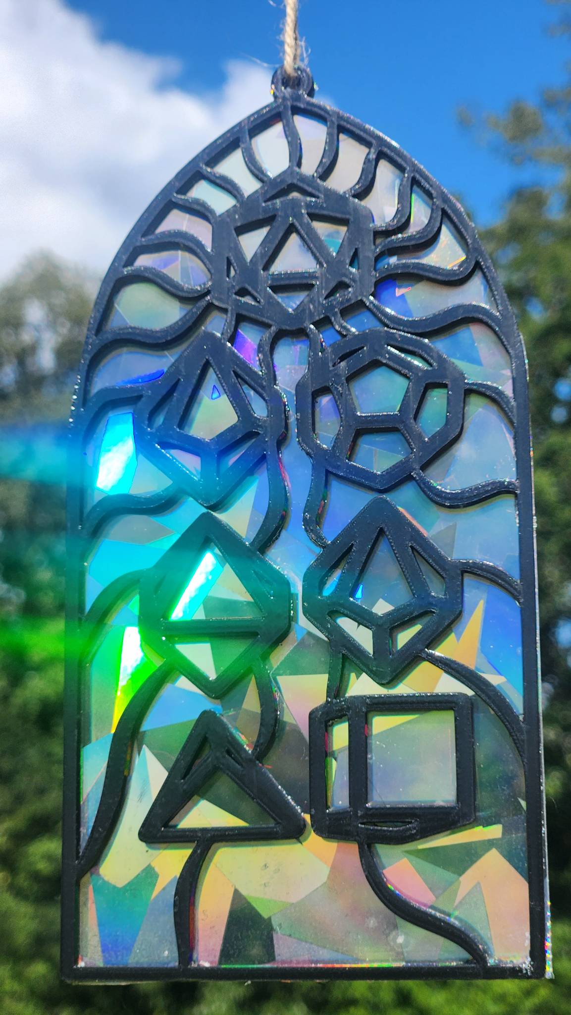 A rainbow film suncatcher with a black filament frame and a dice set, shown outdoors against a blue sky and green trees.