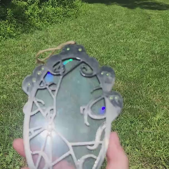A black framed rainbow suncatcher with spooky spirits around a cracked mirror is moved around in the sunlight to showcase colors