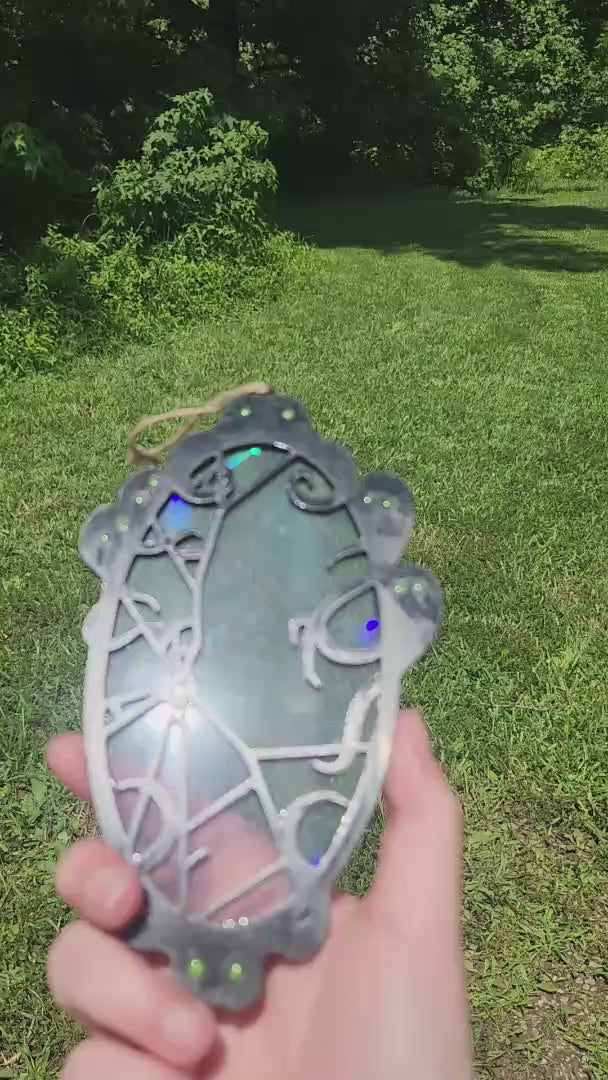 A black framed rainbow suncatcher with spooky spirits around a cracked mirror is moved around in the sunlight to showcase colors