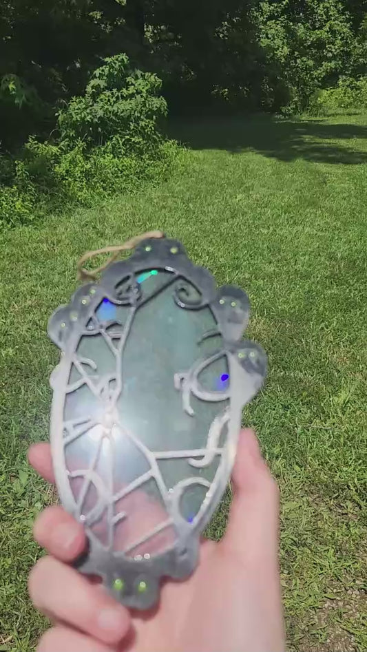 A black framed rainbow suncatcher with spooky spirits around a cracked mirror is moved around in the sunlight to showcase colors