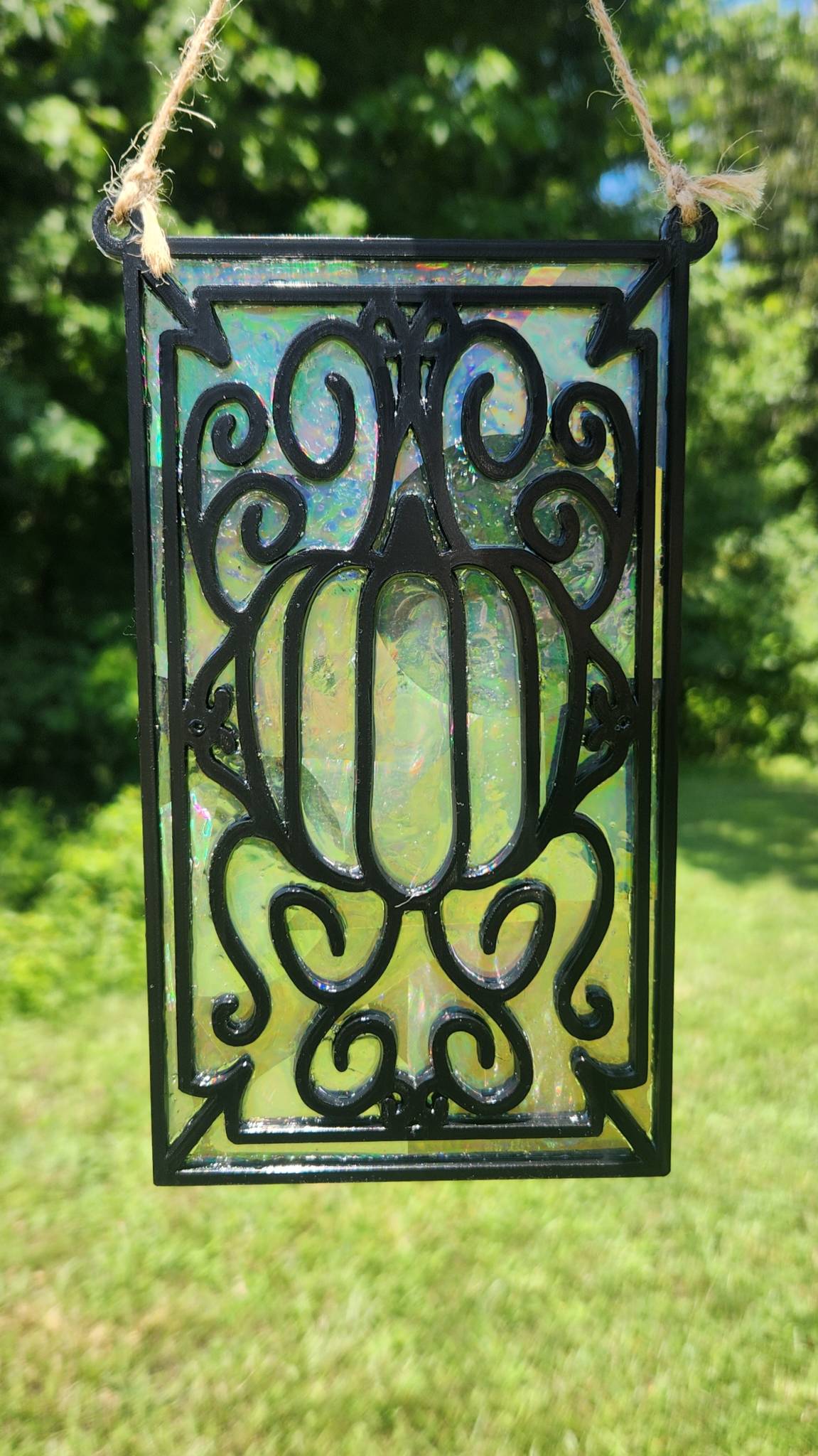 A rainbow suncatcher featuring a pumpkin in the middle of swirls with black filament tied with twine being held outside