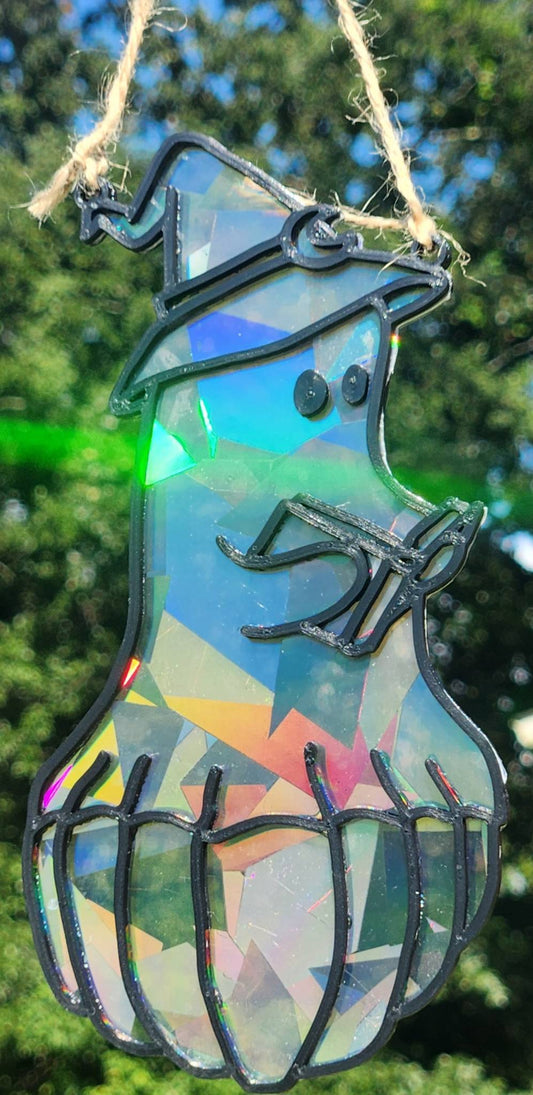 A rainbow suncatcher featuring a ghost sitting on a pumpkin reading hanging outdoors against a blurred green background.