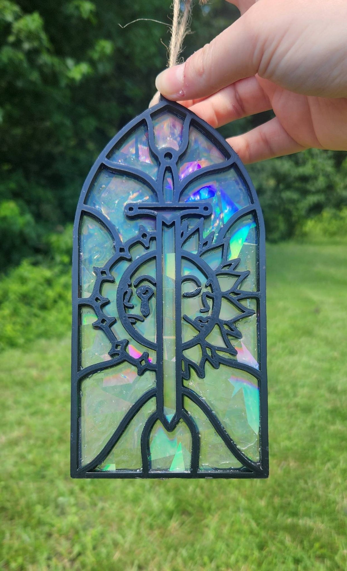 A rainbow suncatcher with black filament outlining featuring a sun and moon split by a sword in a stained glass style held outdoors